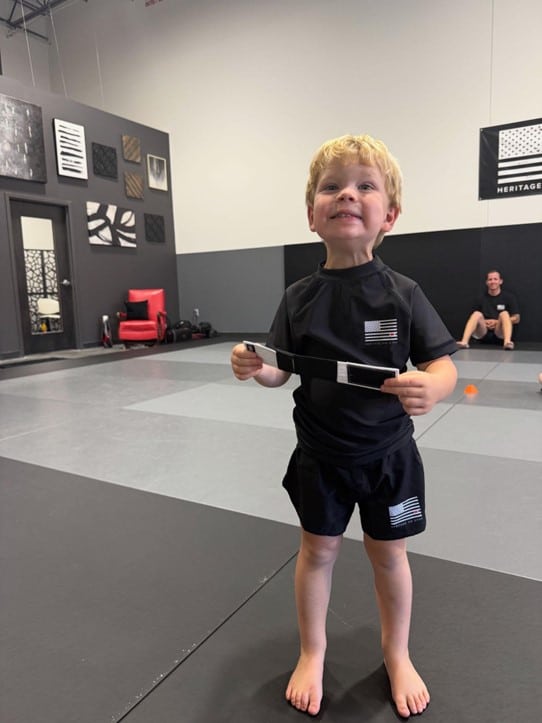 Un niño pequeño sostiene con orgullo su tobillera de rango en Heritage Jiu Jitsu